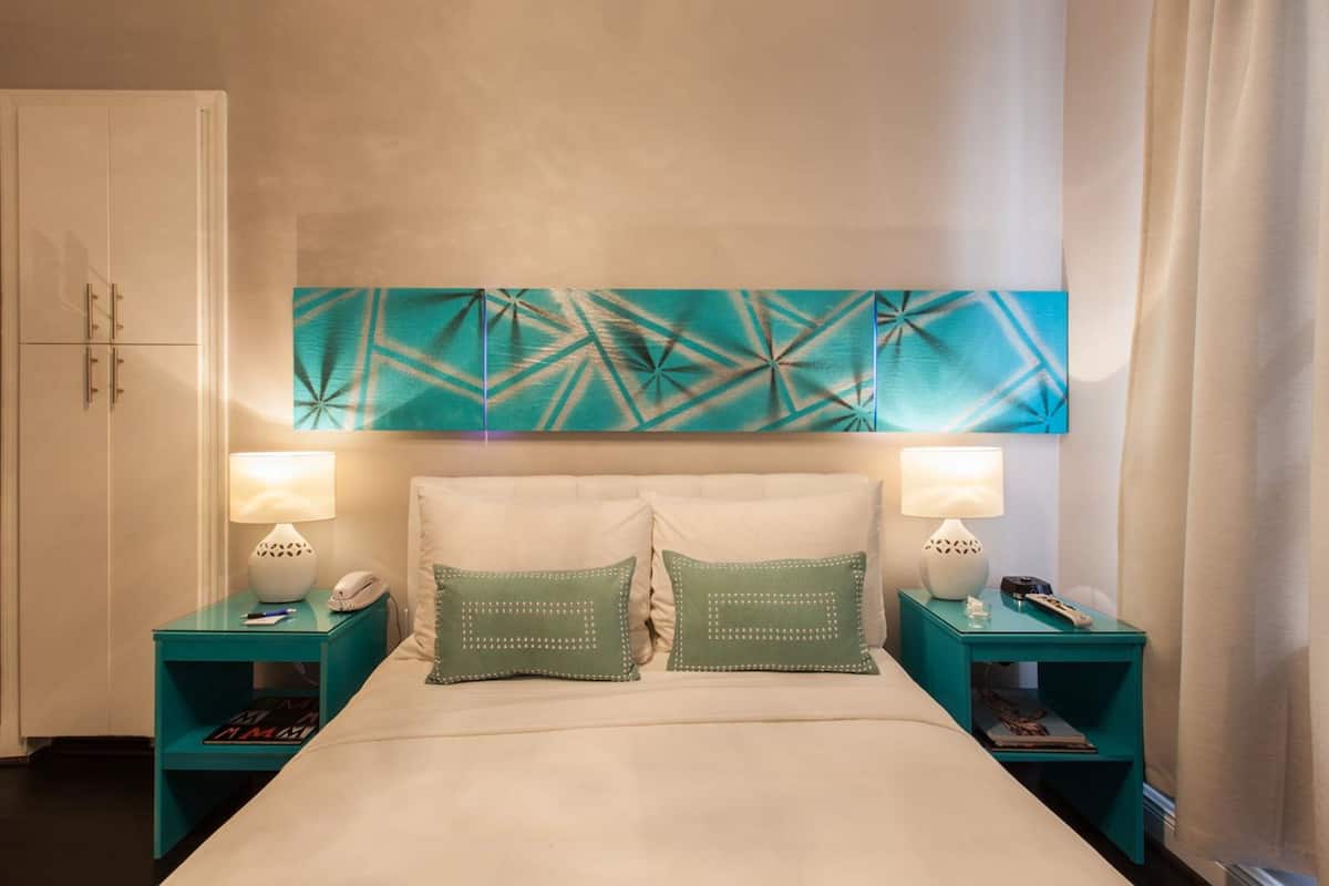 Quarto de hotel com cama, duas mesas de cabeceira, um guarda-roupa branco e uma obra de arte geométrica na parede.