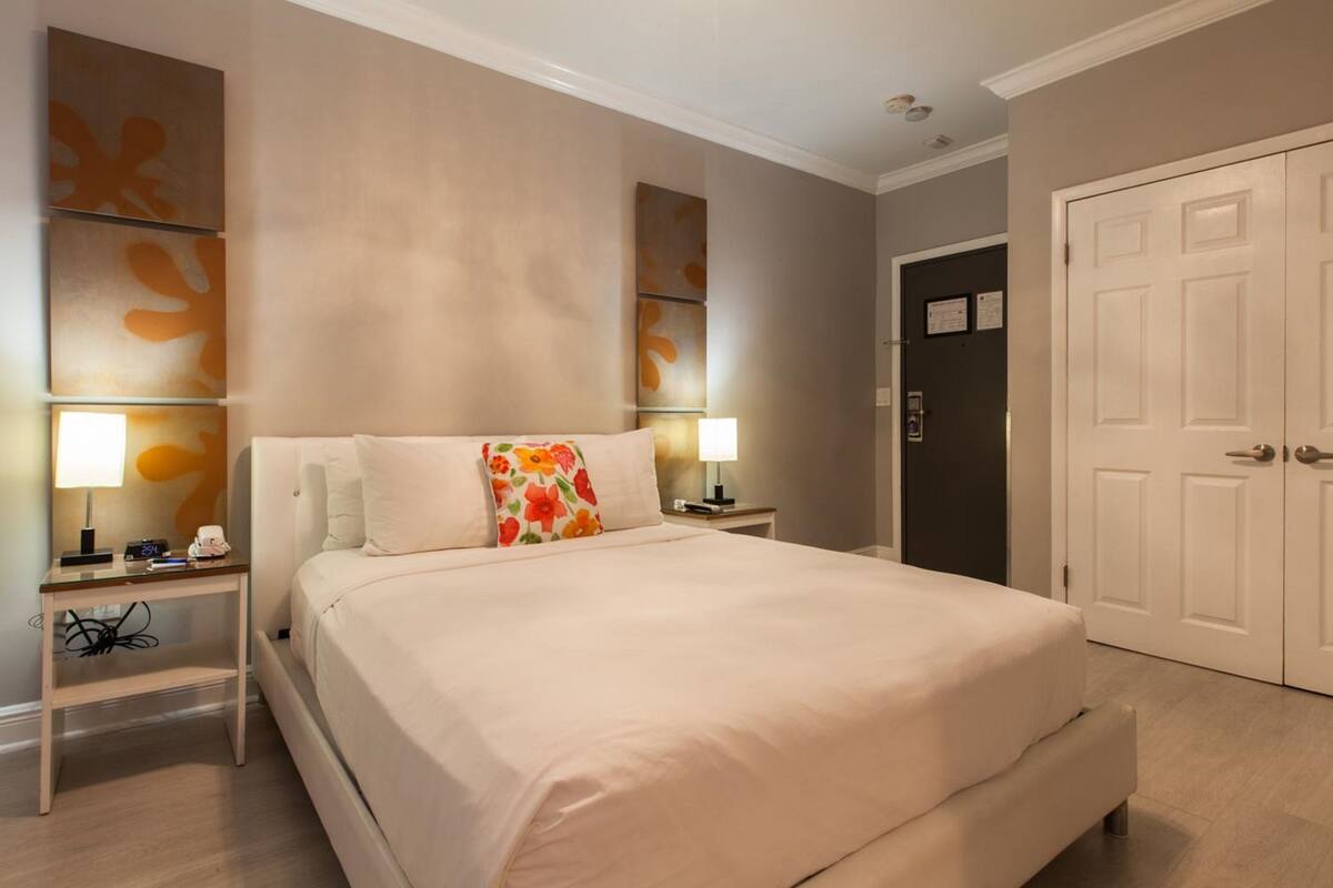 Quarto de hotel com cama, mesas de cabeceira, luminária, porta e parede decorada com arte abstrata.
