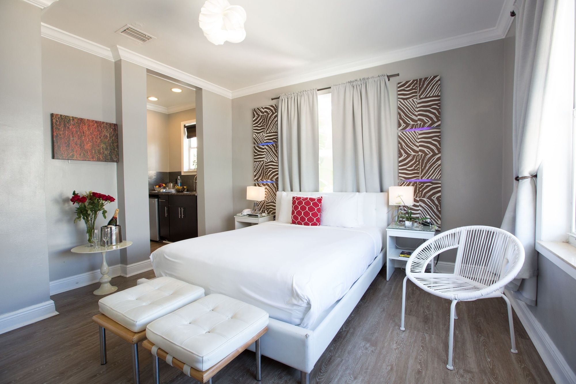 A hotel room with a large bed, bedside tables, a white chair, a small table with flowers, and a view of the kitchen.