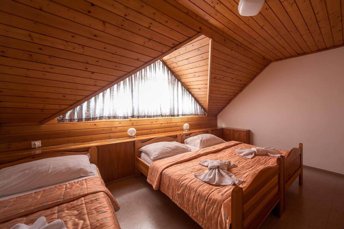 Ein kleines Schlafzimmer im Dachgeschoss mit Holzwänden und schrägen Decken, zwei Betten, einem Fenster mit durchsichtigen Vorhängen und einer einzelnen Lampe.