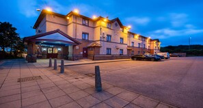Un hotel de varios pisos con un acceso cubierto, zona de estacionamiento y una fila de automóviles estacionados.