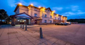 Un hotel de varios pisos con un acceso cubierto, zona de estacionamiento y una fila de automóviles estacionados.