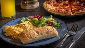 Um prato com batatas fritas, um pão e uma salada, com uma pizza ao fundo.
