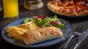 A plate with fries, a bread roll, and a salad, with a pizza in the background.