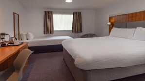 Quarto de hotel com duas camas, uma mesa de trabalho com cafeteira, uma cadeira, uma janela com cortinas e uma luminária na parede.