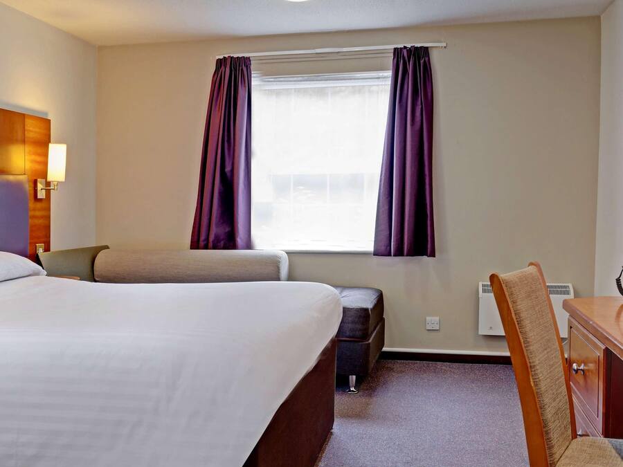 Quarto de hotel com cama, escrivaninha com cadeira, espelho e janela com cortinas.