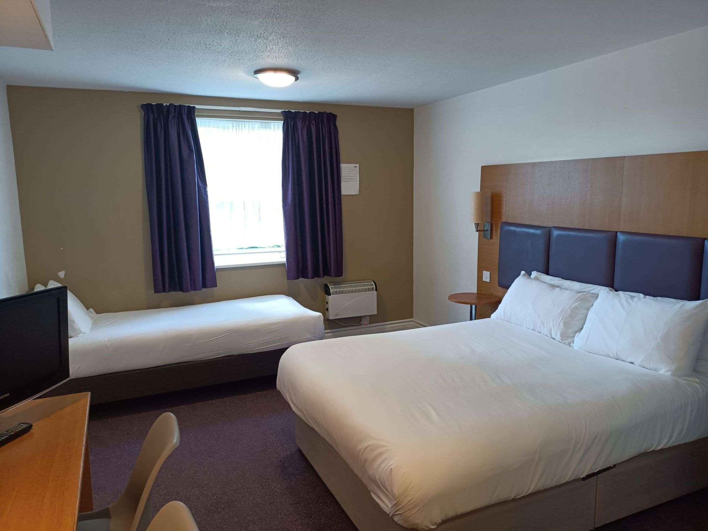 A hotel room with two beds, a desk with a TV, a window with curtains, and a wall-mounted heater.