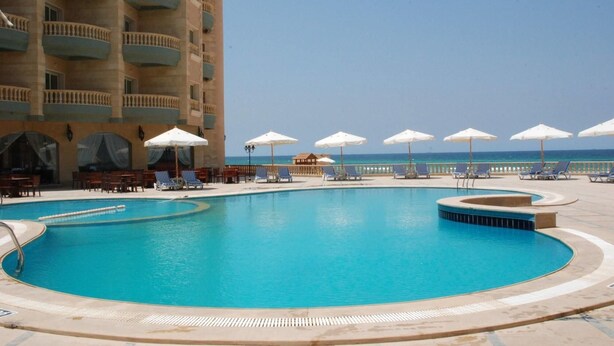 A pool area with lounge chairs and umbrellas, a building with balconies, and a clear view of the ocean. - Royal Sea View (Alexandria)