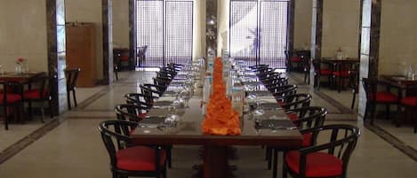 Una larga mesa de comedor con sillas, un adorno naranja en el centro, y vista de un edificio a través de una ventana con diseño.