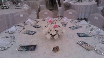 Una sala da banchetti con tavoli rotondi, ognuno apparecchiato con tovaglie bianche, sedie e composizioni floreali.