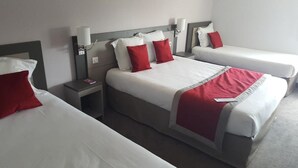A hotel room with two beds, each with white bedding and red pillows, a bedside table with a lamp, and a wall-mounted light fixture.