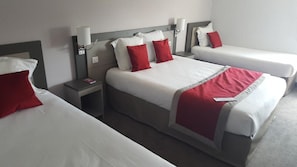 Quarto de hotel com duas camas, cada uma com roupa de cama branca e almofadas vermelhas, mesa de cabeceira com abajur e luminária na parede.