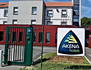 A hotel entrance with a green gate, a sign for Akena Hotels, and a building with a red facade and multiple windows. - Akena City Paris Nord Saint Witz (Saint-Witz)