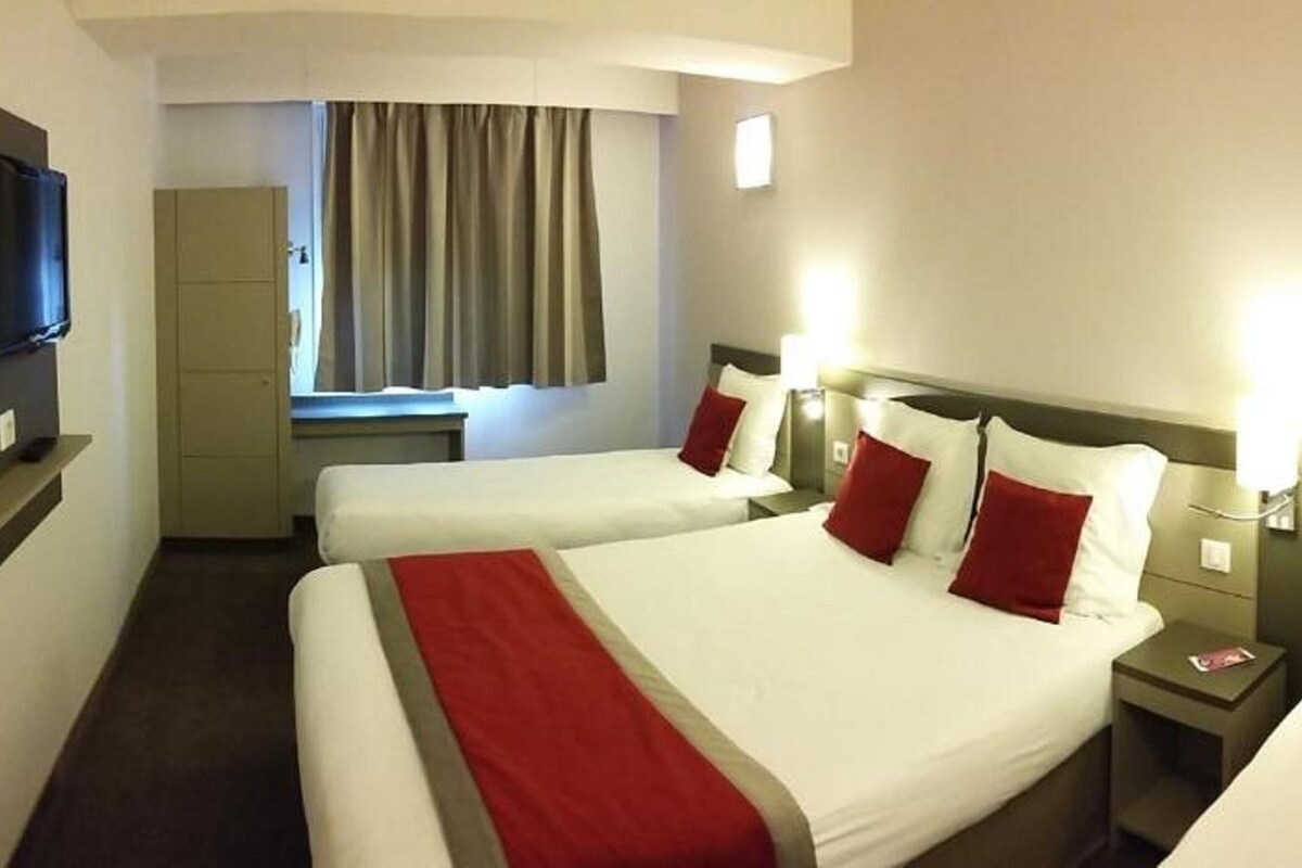 Une chambre d’hôtel avec trois lits, une télévision, une fenêtre avec des rideaux et des lampes de chevet.