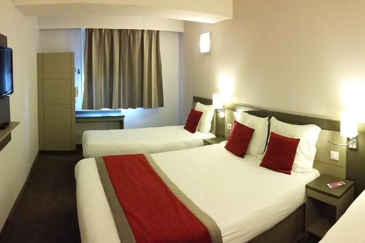A hotel room with three beds, a television, a window with curtains, and bedside lamps.
