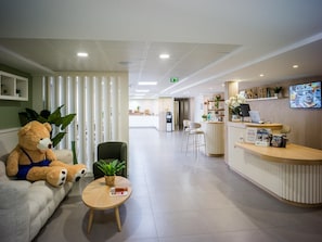 A hotel reception area with a reception desk, a seating area with a sofa and a chair, a table with a plant, and a large teddy bear.