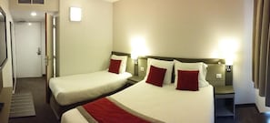 Quarto de hotel com duas camas, uma mesinha de cabeceira, uma luminária e uma porta.