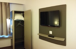 A hotel room with a television mounted on the wall, a closet with a hanging garment, and a window with curtains.