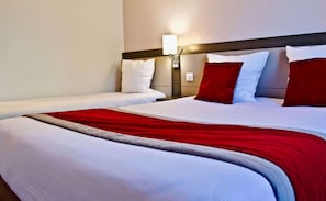 Quarto de hotel com duas camas, uma mesa de cabeceira com abajur e uma colcha vermelha e branca.