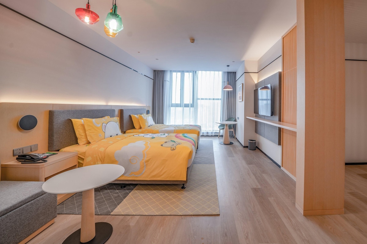 A modern hotel room with a large bed, bedside tables, a desk, a TV, and a children's play area.