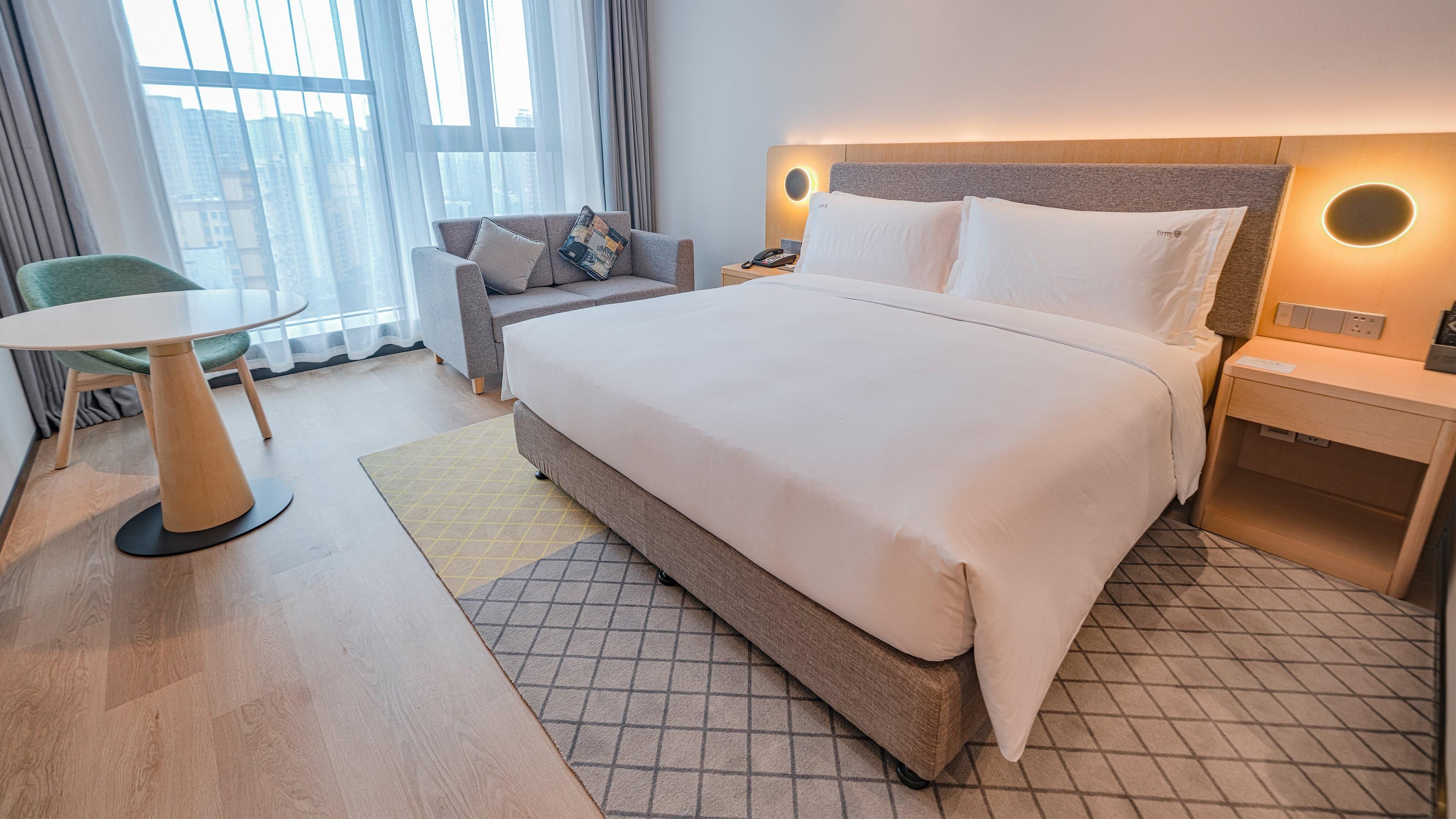 A hotel room with a large bed, a bedside table with a lamp, a chair, a small table, and a view of the city through the window.