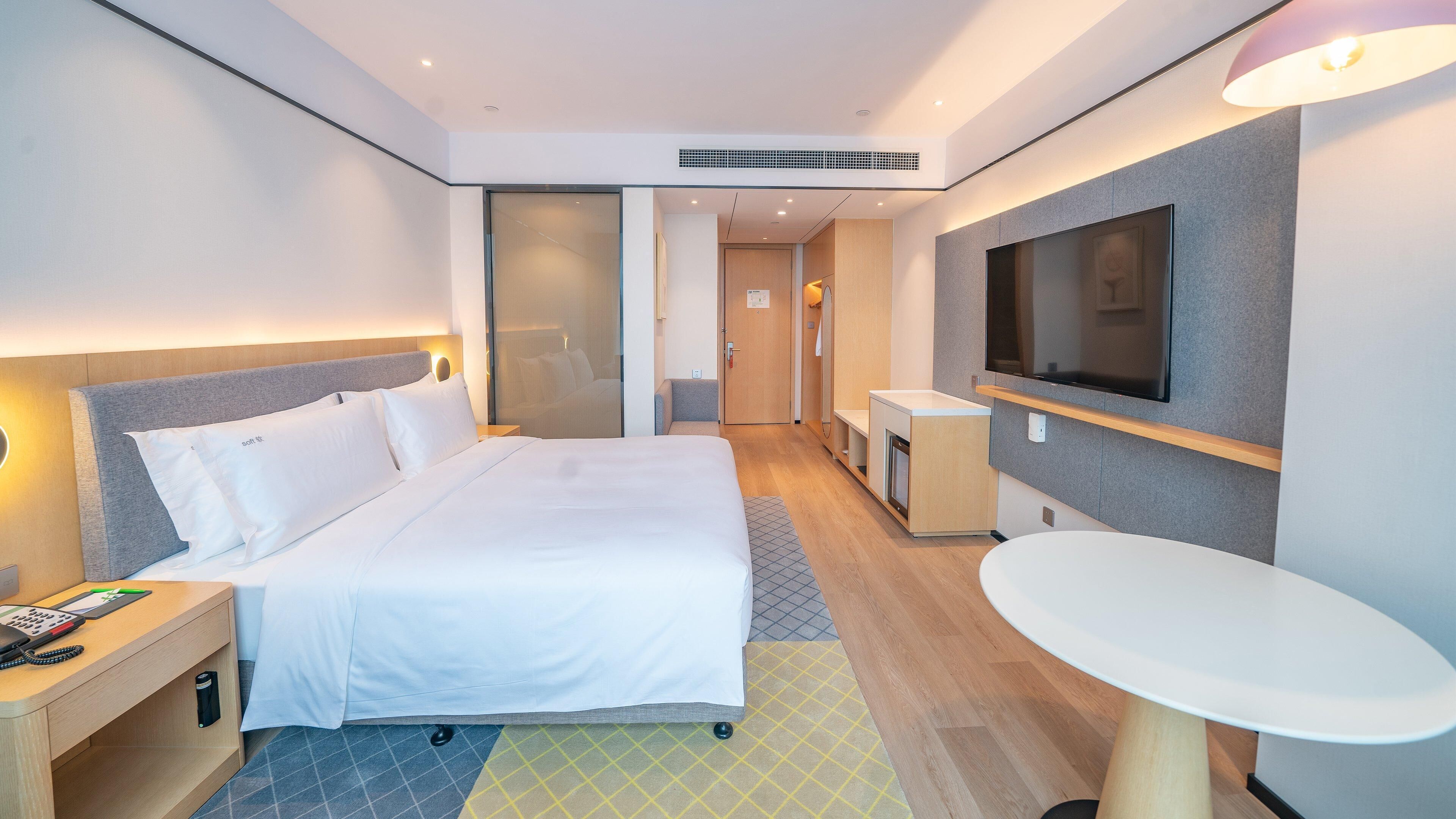 A modern hotel room with a large bed, bedside table with a phone, a small desk, a TV mounted on the wall, and a small round table.