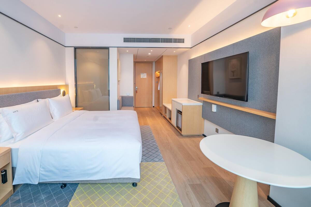 A modern hotel room with a large bed, bedside table with a phone, a small desk, a TV mounted on the wall, and a small round table.