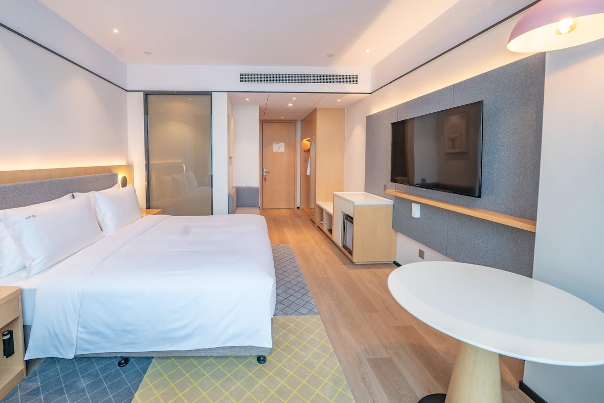 Quarto de hotel moderno com uma cama grande, mesa de cabeceira com telefone, uma pequena escrivaninha, televisão na parede e uma mesa redonda.