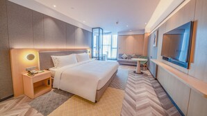 A modern hotel room with a large bed, a bedside table with a phone, a small round table, a sofa, and a TV.