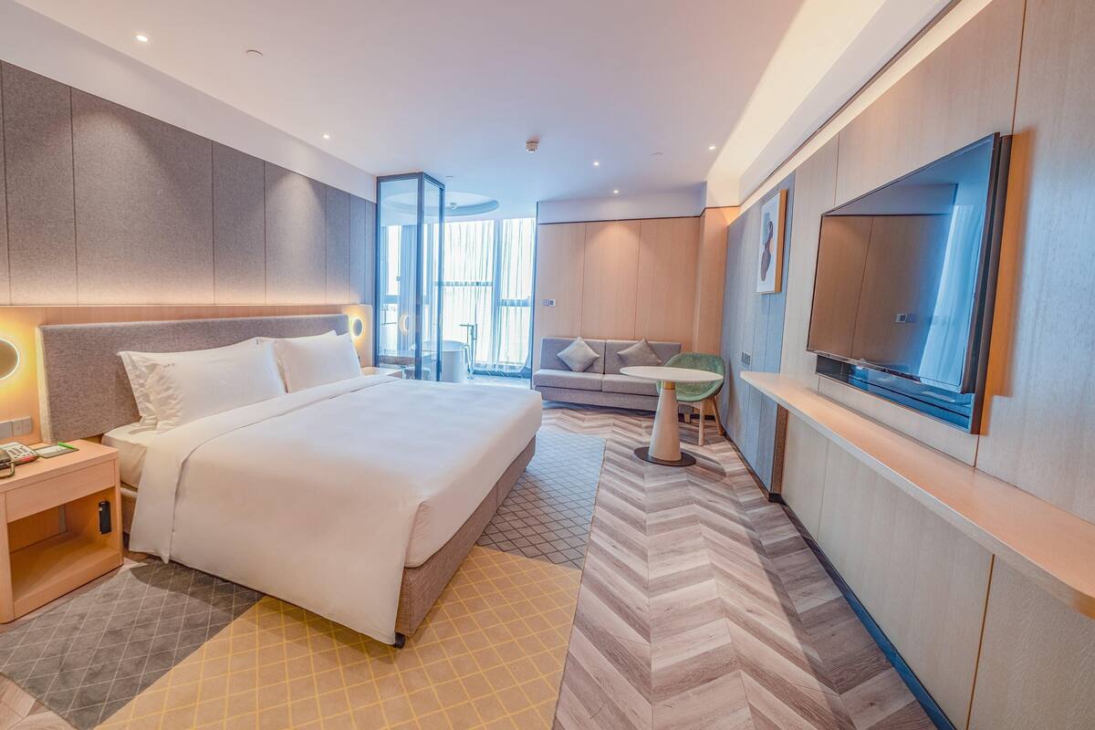 A modern hotel room with a large bed, a bedside table with a phone, a small round table, a sofa, and a TV.