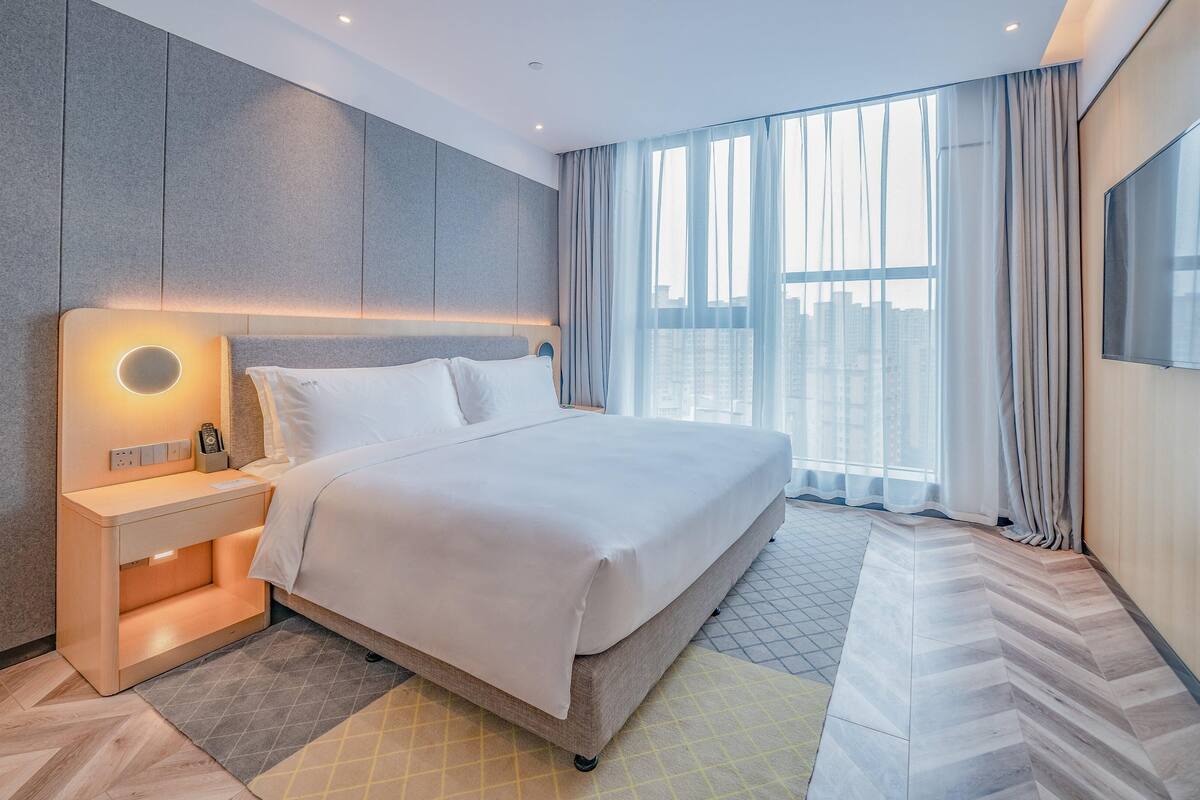 A modern hotel room with a large bed, a bedside table with a lamp, a flat-screen TV mounted on the wall, and a view of the city through large windows.