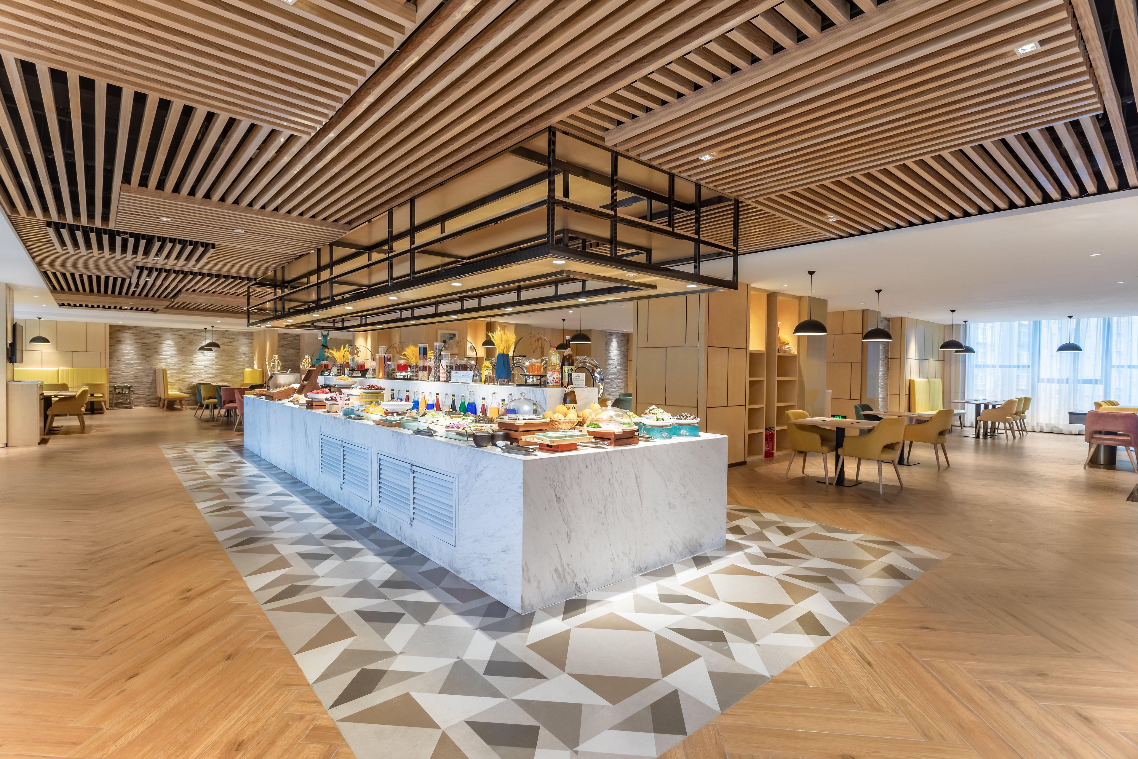 A spacious hotel lobby with a central buffet, wooden ceiling design, and modern furniture.