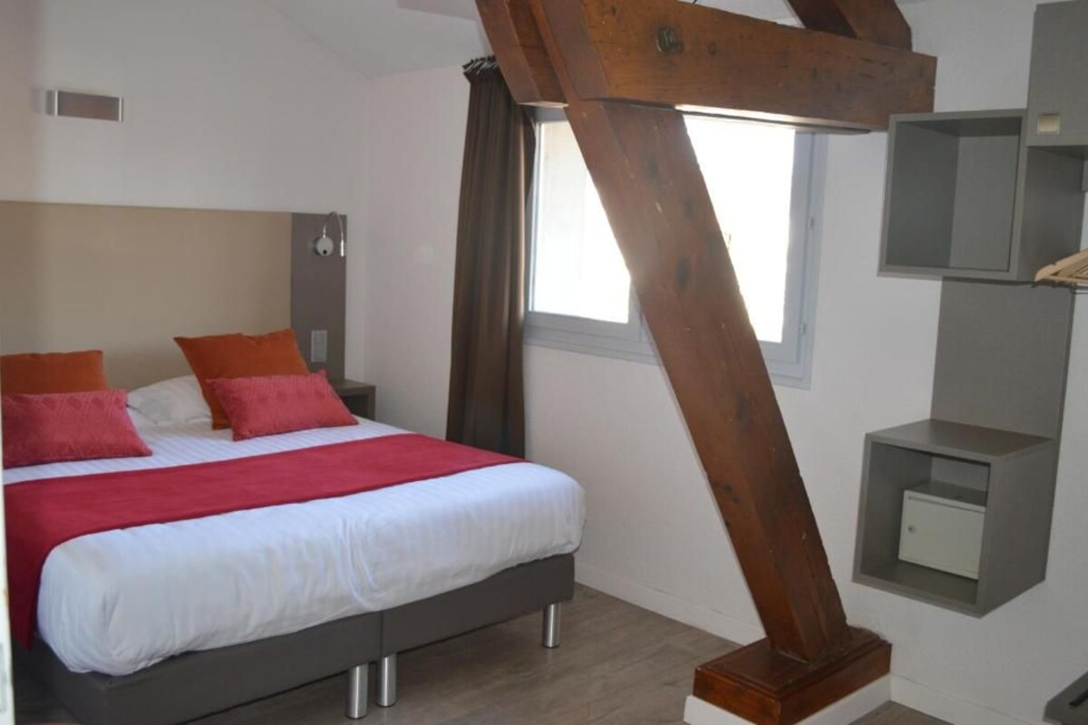 A bedroom with a bed, a nightstand, a wooden beam, a window, and a built-in shelf.
