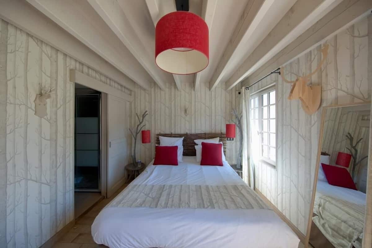 A bedroom with a large bed, a wooden ceiling, a red lamp, and a mirror with a deer head.