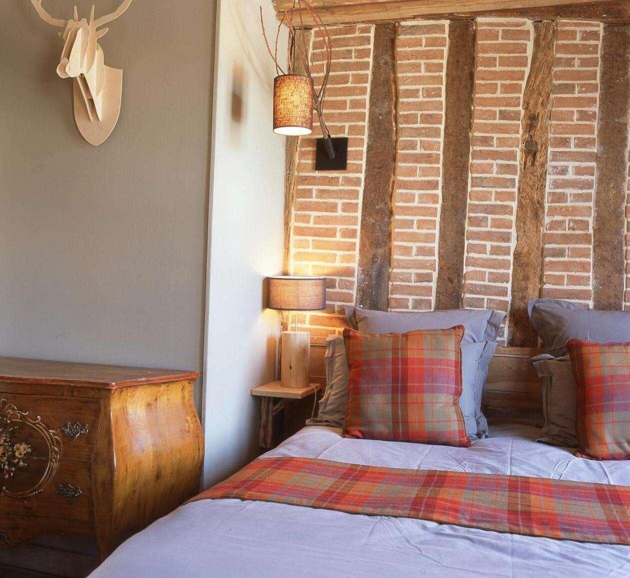 A bedroom with a brick wall, a wooden dresser, a bed with plaid bedding, and a hanging lamp.