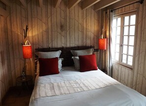 Een slaapkamer met een houten wandpaneel, een bed met witte beddengoed en rode kussens, twee wandlampen en een raam met gordijnen.