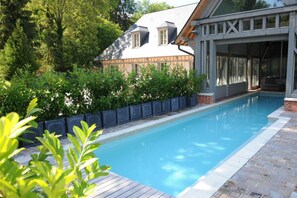 A swimming pool with a wooden deck, surrounded by greenery and a building with a gabled roof. - Hôtel Le Grand Cerf (Lyons-la-Foret)