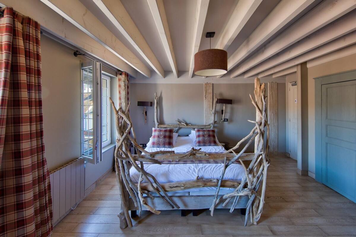 A room with a bed made of branches, a window with curtains, a hanging light fixture, and a door.