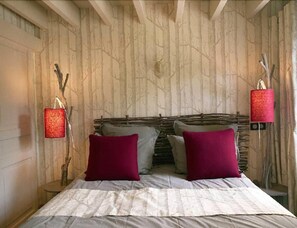 Un dormitorio con cabecera de madera, dos apliques rojos en la pared y una cama con ropa de cama blanca y dos almohadas moradas.