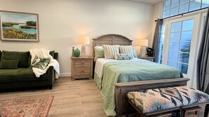 Un dormitorio con una cama, un sofá verde, un banco, una mesita de noche, una lámpara, un cuadro y una ventana.