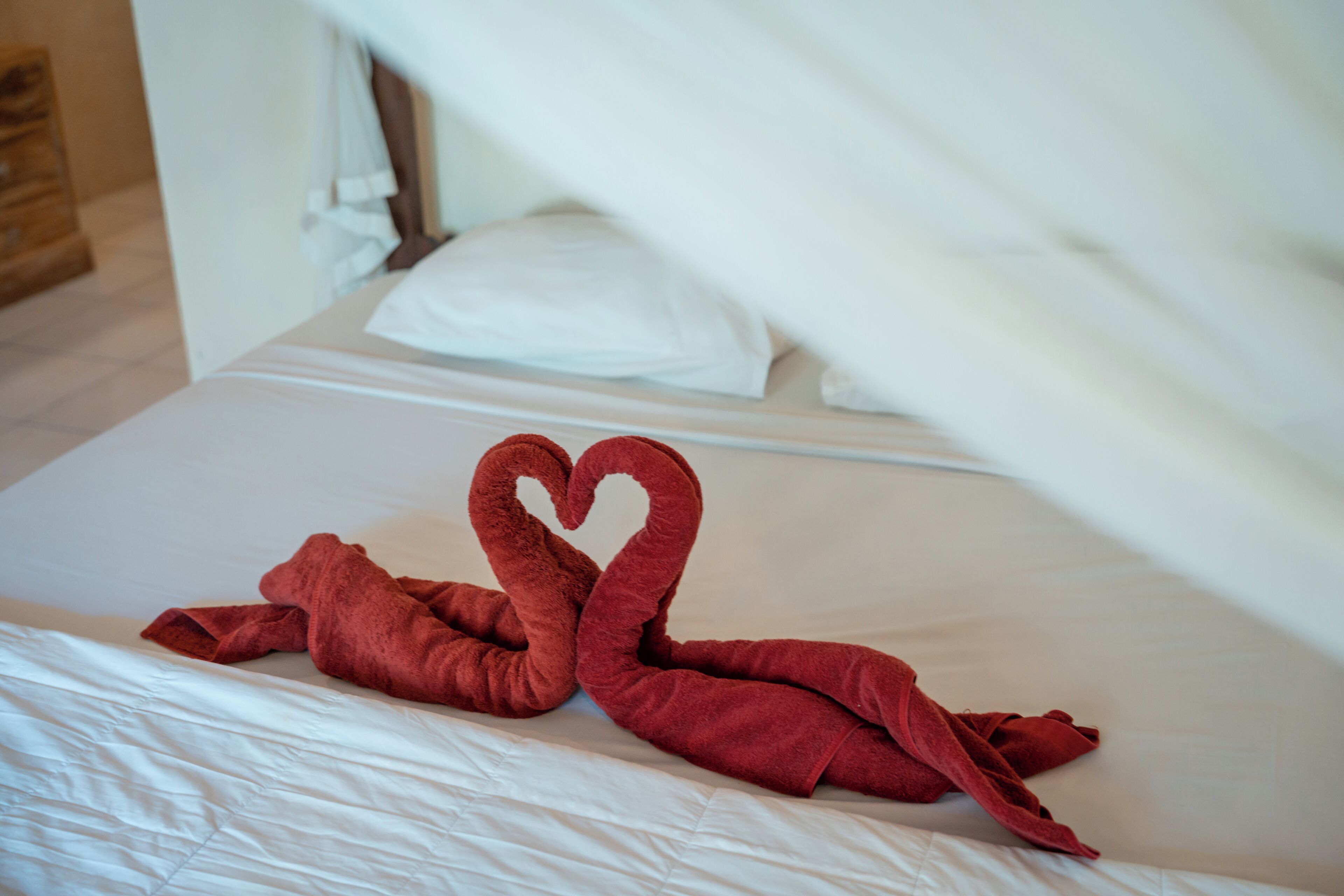 A bed with white linens and a red towel arranged in a heart shape.