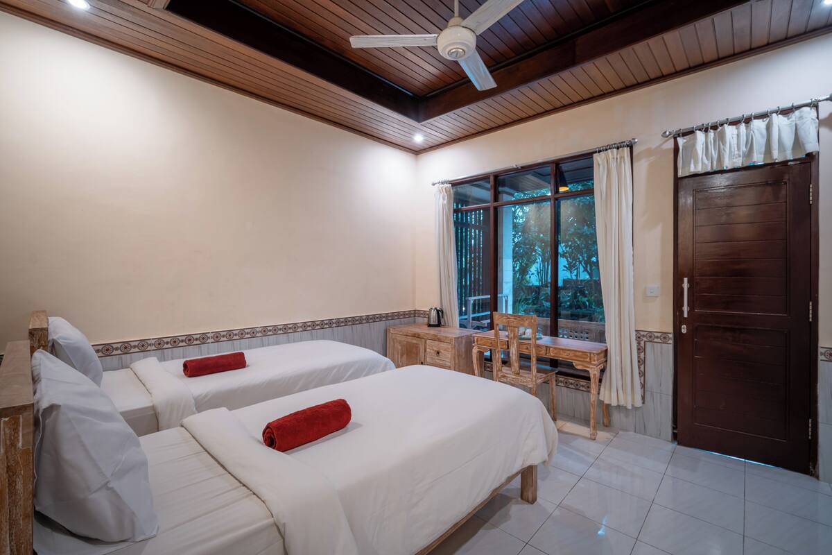 A hotel room with two beds, a wooden desk, a chair, a window with curtains, and a ceiling fan.