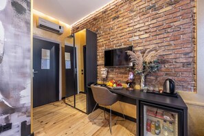 Une chambre d’hôtel avec un mur en briques, un téléviseur à écran plat, un plancher en bois, un bureau avec une chaise, un miroir, un réfrigérateur et un climatiseur fixé au mur.