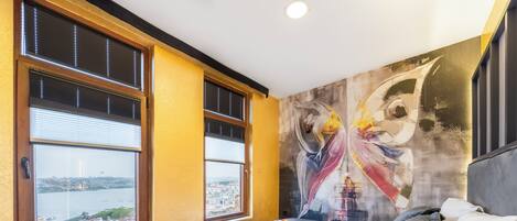Un dormitorio con cama, ventana con vista a la ciudad y un cuadro en la pared.