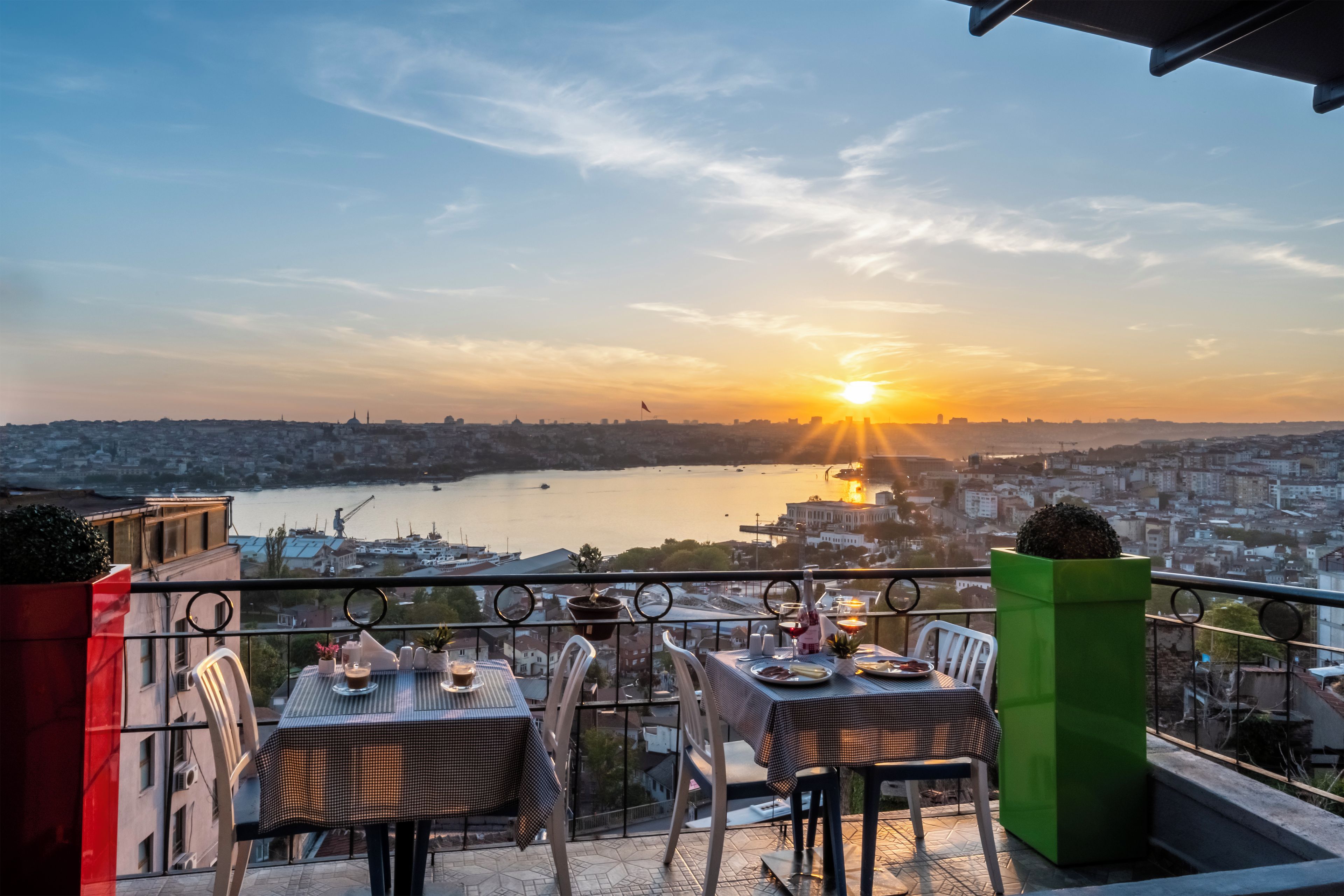 Terraza con mesas y sillas que da a una vista panorámica de la ciudad y un cuerpo de agua al atardecer.