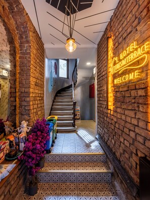 Un hall d’hôtel avec un mur en briques, un panneau lumineux au néon, un escalier et un comptoir d’accueil.