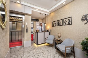 Un hall d’hôtel comprenant un comptoir d’accueil, deux fauteuils, un sol à motifs et un mur orné de tableaux encadrés.