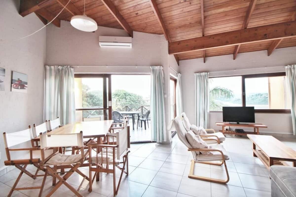 Une pièce spacieuse avec un plafond en bois, une table à manger avec des chaises, un canapé, un téléviseur et de grandes fenêtres avec des rideaux.