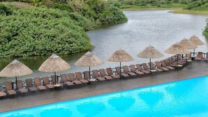 A pool with lounge chairs and umbrellas, surrounded by lush greenery and a river. - Top Destinations (Southbroom)