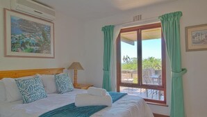A hotel room with a bed, a window with curtains, a painting on the wall, and an air conditioning unit. - Top Destinations (Southbroom)